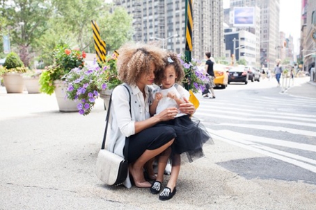 A biracial mother is crouching and hugging her daughter near a city street with tall buildings.